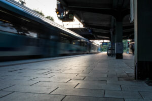 Eine verschwommene Darstellung eines Zuges, der schnell an einem Bahnhof vorbeifährt. Der Blick ist von unten auf den Bahnhof gerichtet, wobei die hellen Fliesen des Bahnsteigs hervorgehoben werden. Im Hintergrund sind Elemente wie ein Bahnhofsschild und Verkehrssignale sichtbar, und die Szene vermittelt ein Gefühl von Bewegung und
