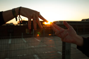 Zwei Hände, die sich in der Abenddämmerung strecken, wobei die Fingerspitzen fast die untergehende Sonne berühren. Im Hintergrund sind unscharfe Konturen von Gebäuden und einem Zaun sichtbar, während die Atmosphäre eine warme und friedliche Stimmung vermittelt.